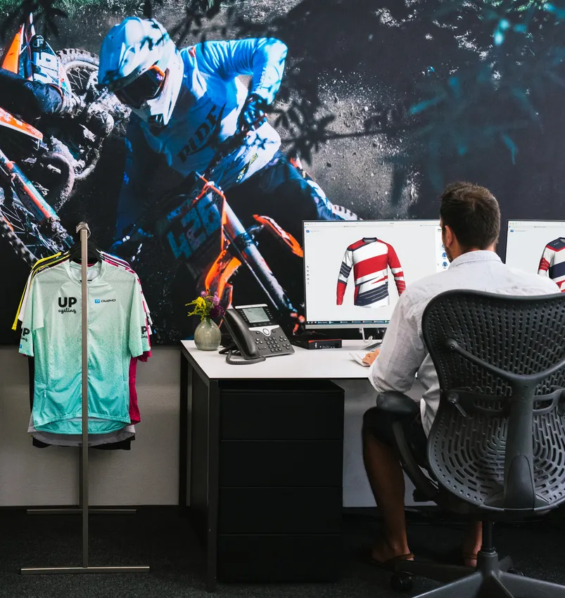 Un uomo è seduto a una scrivania e progetta su un computer una maglia sportiva personalizzata, mentre sullo sfondo si vede una grande immagine di un motociclista e un portabiti con vari indumenti sportivi.