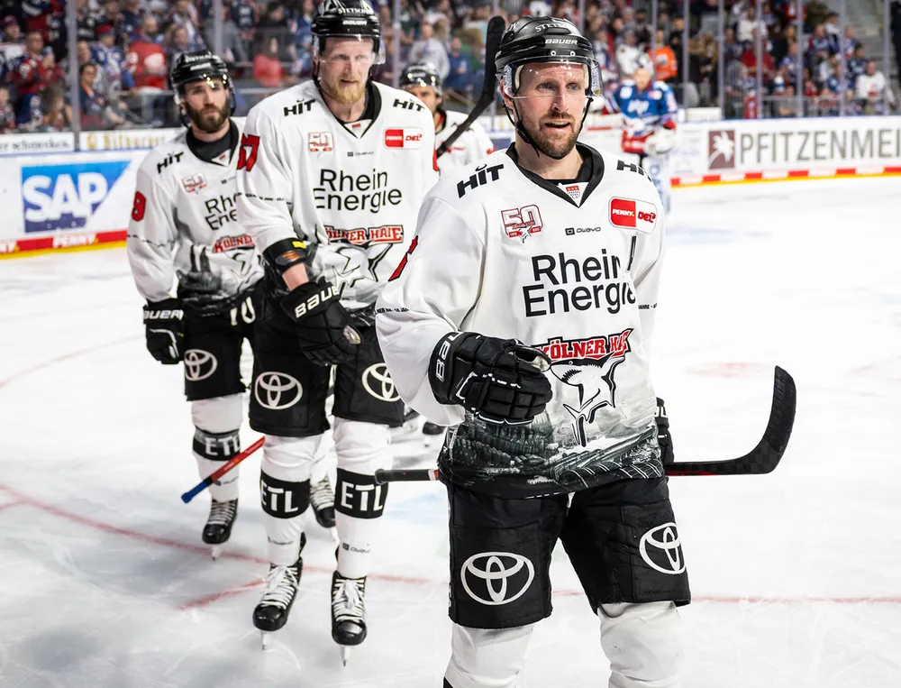 Tre giocatori di hockey su ghiaccio in maglie bianche con loghi neri e design grafici stanno in piedi sul ghiaccio.
