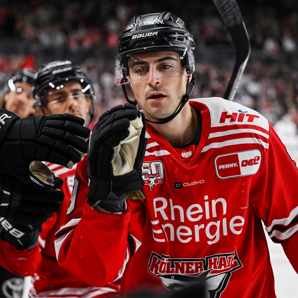 Un giocatore di hockey in una maglia rossa con strisce bianche e loghi, in piedi sul ghiaccio e sollevando i guanti.