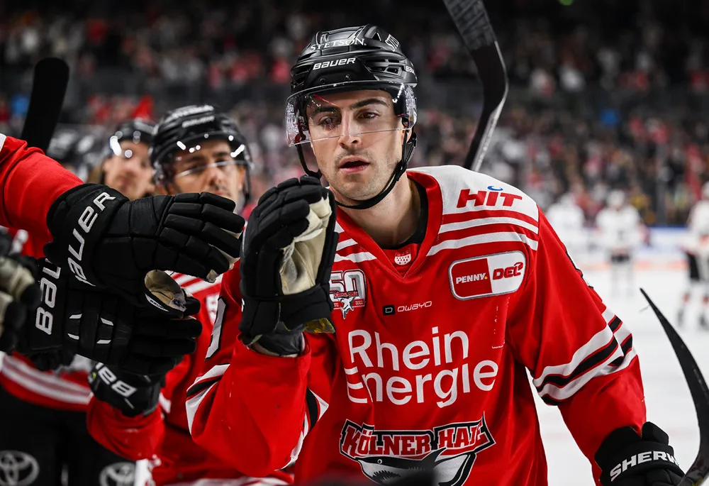Un giocatore di hockey su ghiaccio in maglia rossa con guanti neri e casco, che festeggia con i compagni di squadra.
