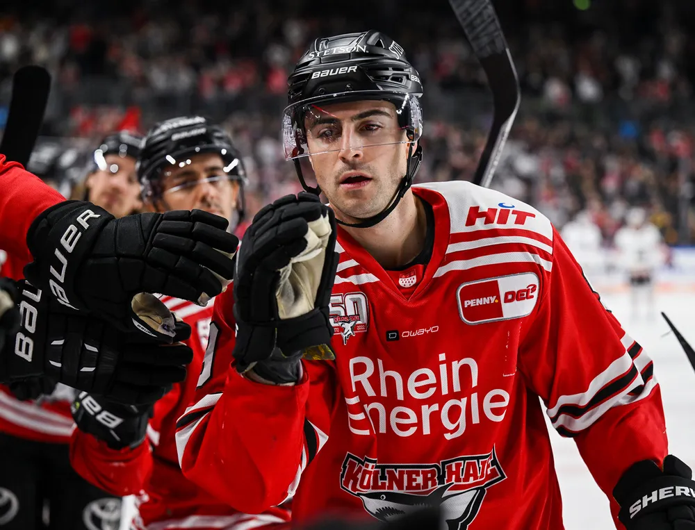 Un giocatore di hockey indossa una maglia rossa con dettagli bianchi e neri mentre interagisce con un compagno di squadra.