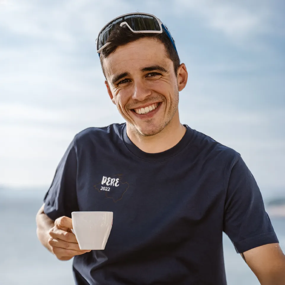Un uomo tiene una tazza in mano e indossa una maglietta blu scuro con la stampa 'PERE 2022', sorridendo davanti a uno sfondo sfocato.