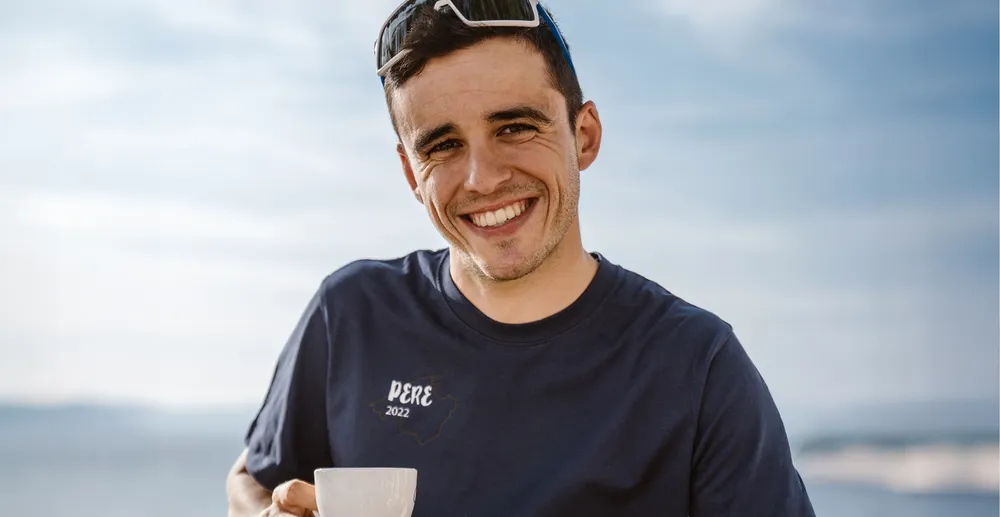 Un uomo sportivo con una maglietta blu navy con la scritta 'PERE 2022' sorride mentre tiene una tazza, sullo sfondo di un paesaggio costiero sfocato.