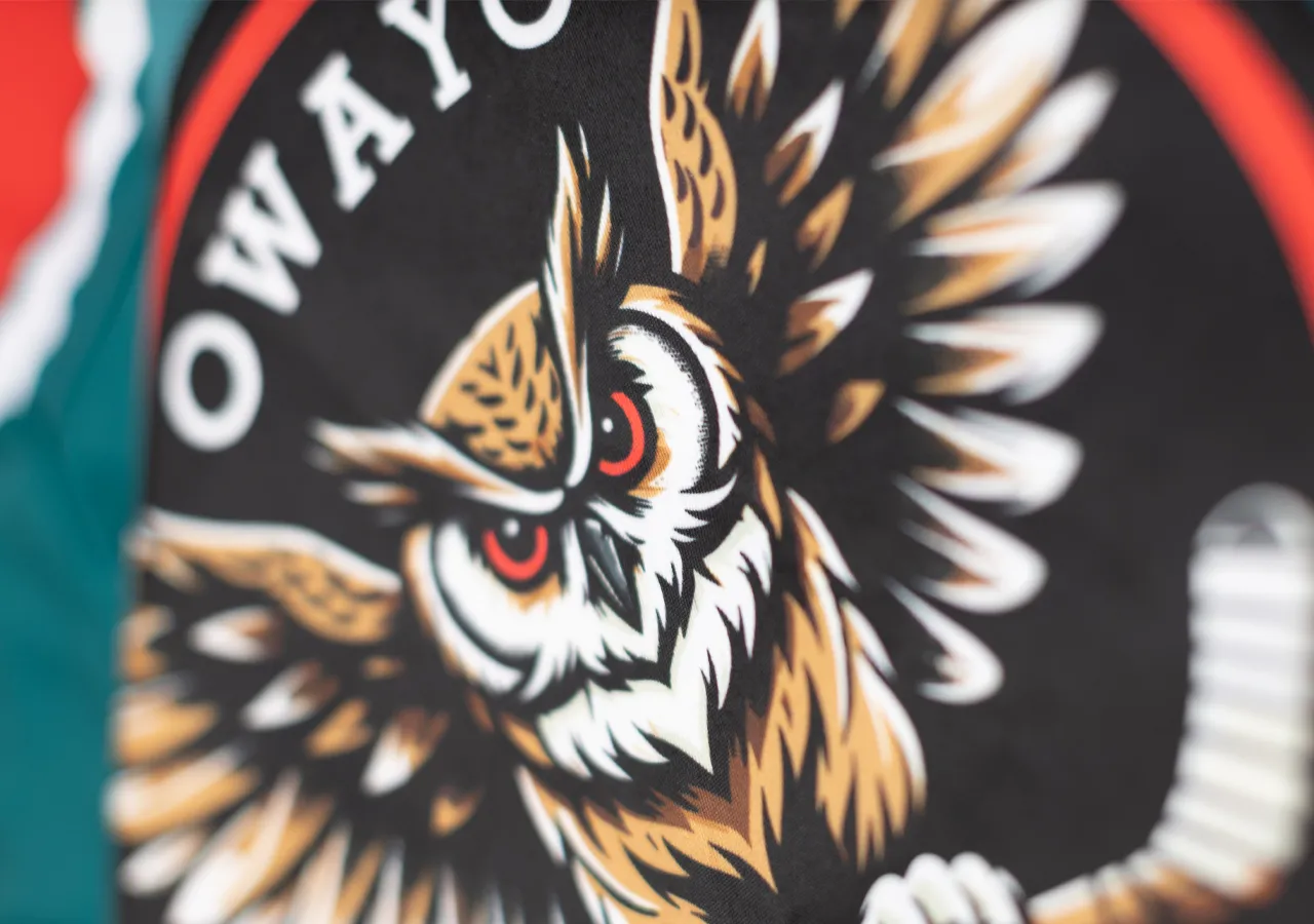 Una testa di gufo stilizzata con occhi rossi su sfondo nero, circondata da un cerchio rosso e bianco, su un capo di abbigliamento sportivo di owayo.
