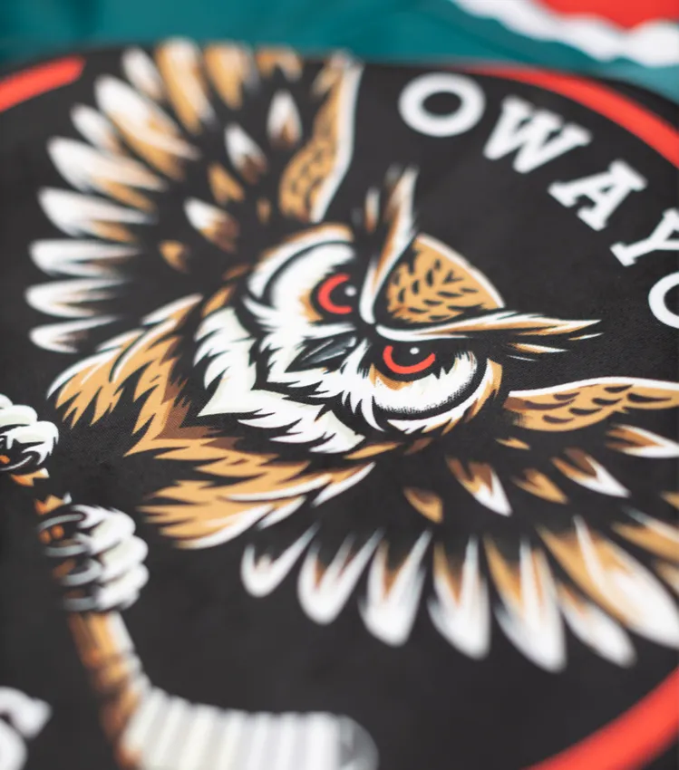 Un primo piano di una maglia sportiva nera con un logo stilizzato di gufo che tiene una stecca da hockey, e la scritta 'owayo' in caratteri bianchi.