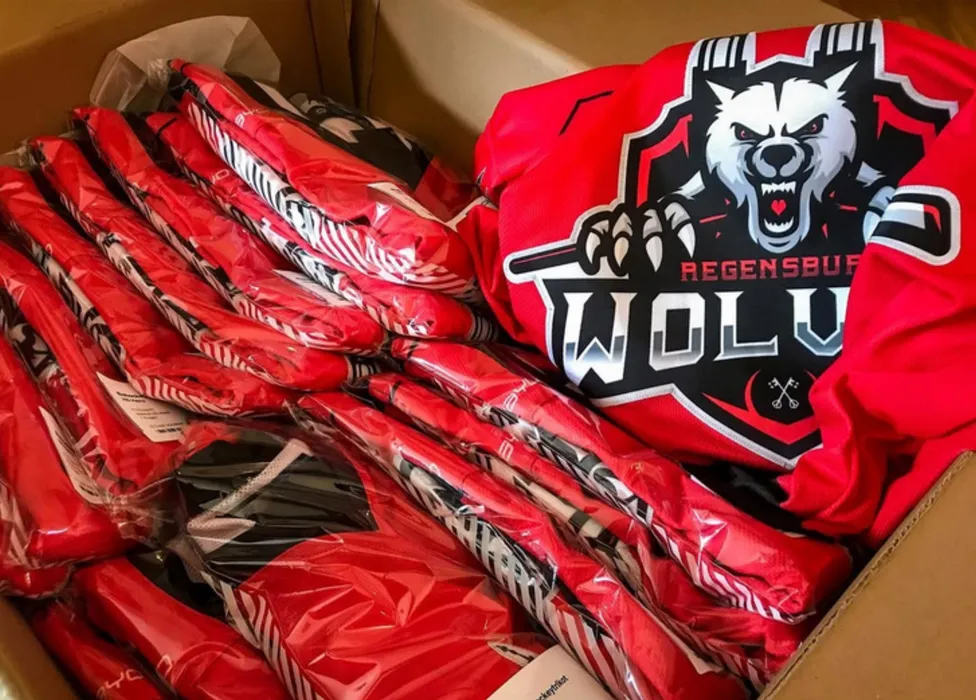 Una scatola piena di maglie sportive rosse personalizzate, tra cui una con un logo di lupo e la scritta 'Regensburg Wolves'.