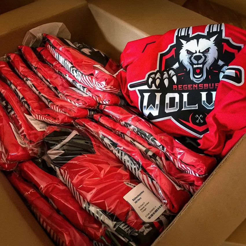 Una scatola piena di diverse magliette sportive rosse avvolte in plastica, inclusa una maglietta visibile con un logo di lupo e il testo 'Regensburg WOLV'.