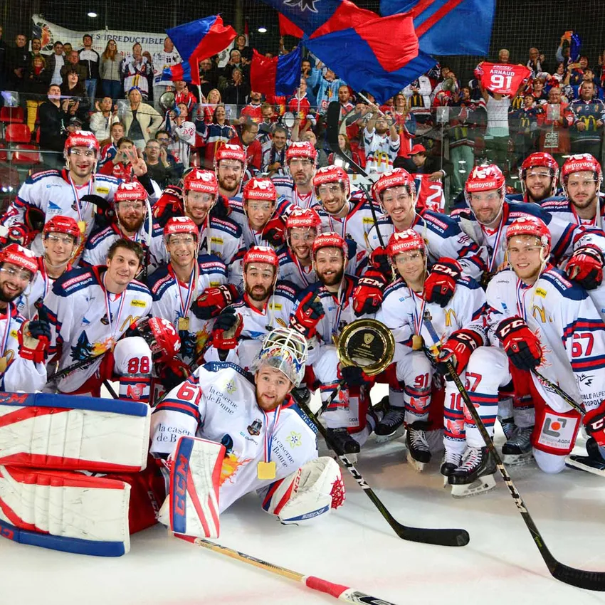 Un gruppo di giocatori di hockey su ghiaccio in maglie bianche con accenti rossi e blu posa orgogliosamente con medaglie e un trofeo sul ghiaccio.