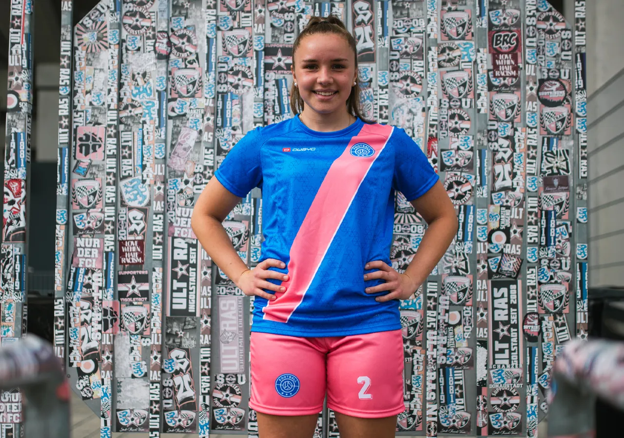Una giovane donna indossa una maglietta da calcio blu con una striscia diagonale rosa e pantaloni corti rosa con il numero 2, davanti a un muro coperto di grafiche e adesivi.
