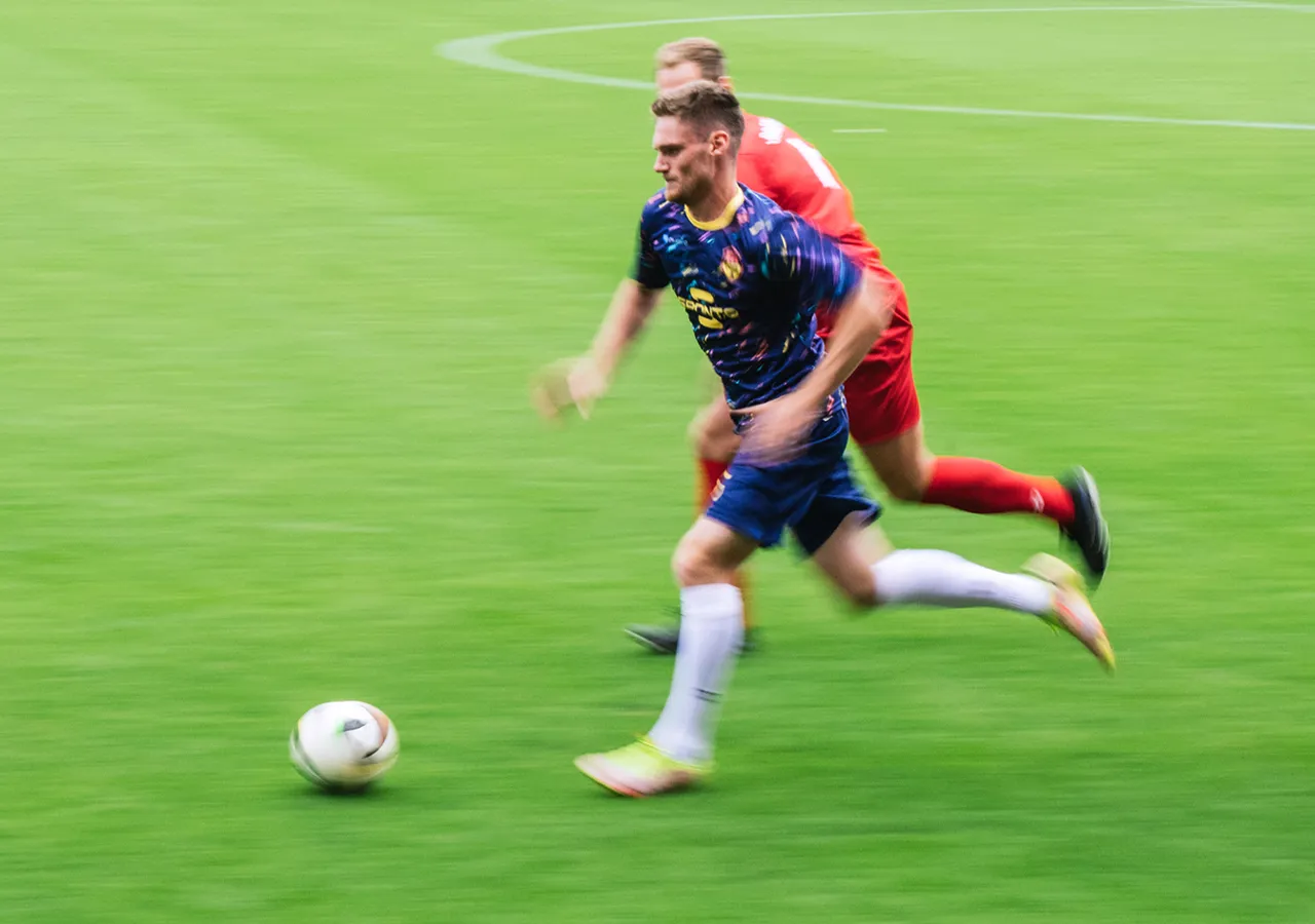 Un calciatore in una maglietta colorata corre sull'erba mentre un altro giocatore lo segue da vicino.