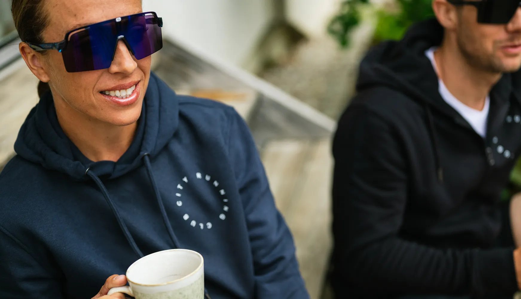 Una persona con un hoodie blu tiene in mano una tazza e indossa grandi occhiali da sole, mentre un'altra persona sullo sfondo indossa anch'essa un hoodie.