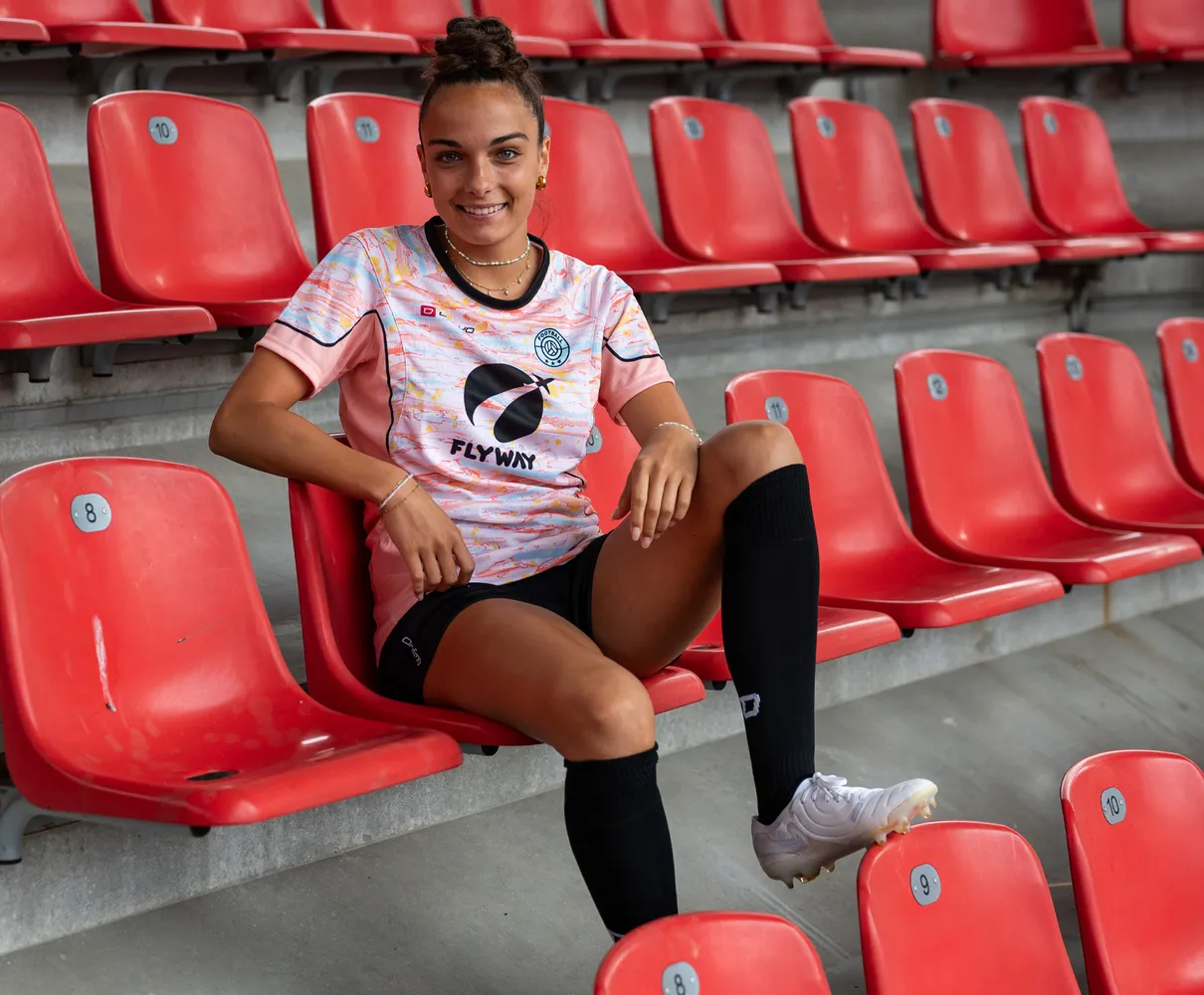 Una giovane donna è seduta su una sedia rossa dello stadio, indossando una maglietta da calcio colorata e calze nere.