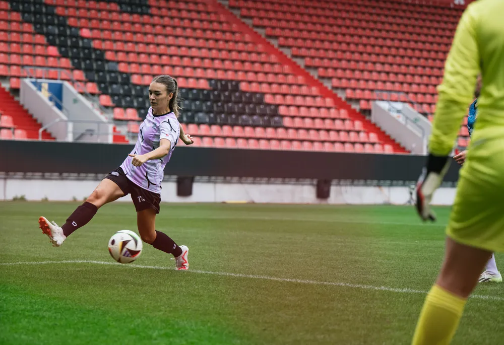 Una calciatrice in maglia viola e nera calcia un pallone su un campo con tribune vuote sullo sfondo.
