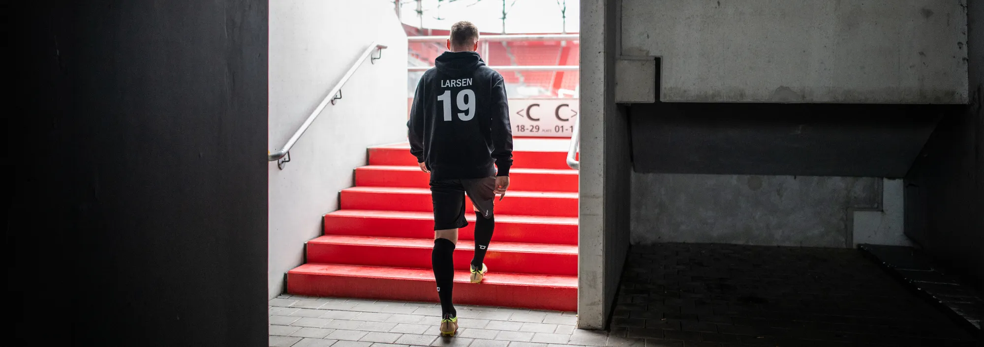 Un giocatore con un felpa scura e pantaloni corti sta salendo le scale rosse di uno stadio.