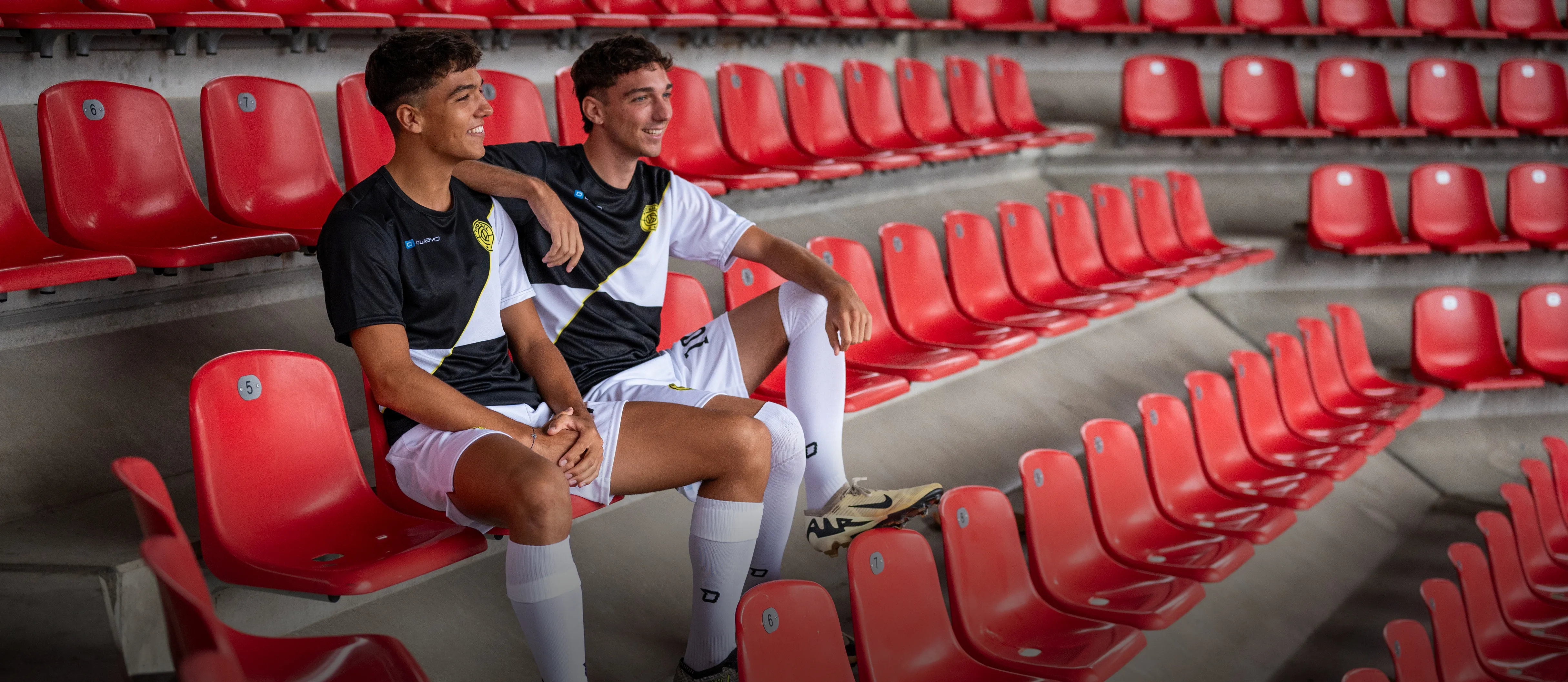 Due giovani in abbigliamento sportivo da calcio sono seduti in modo informale su seggiolini rossi dello stadio.