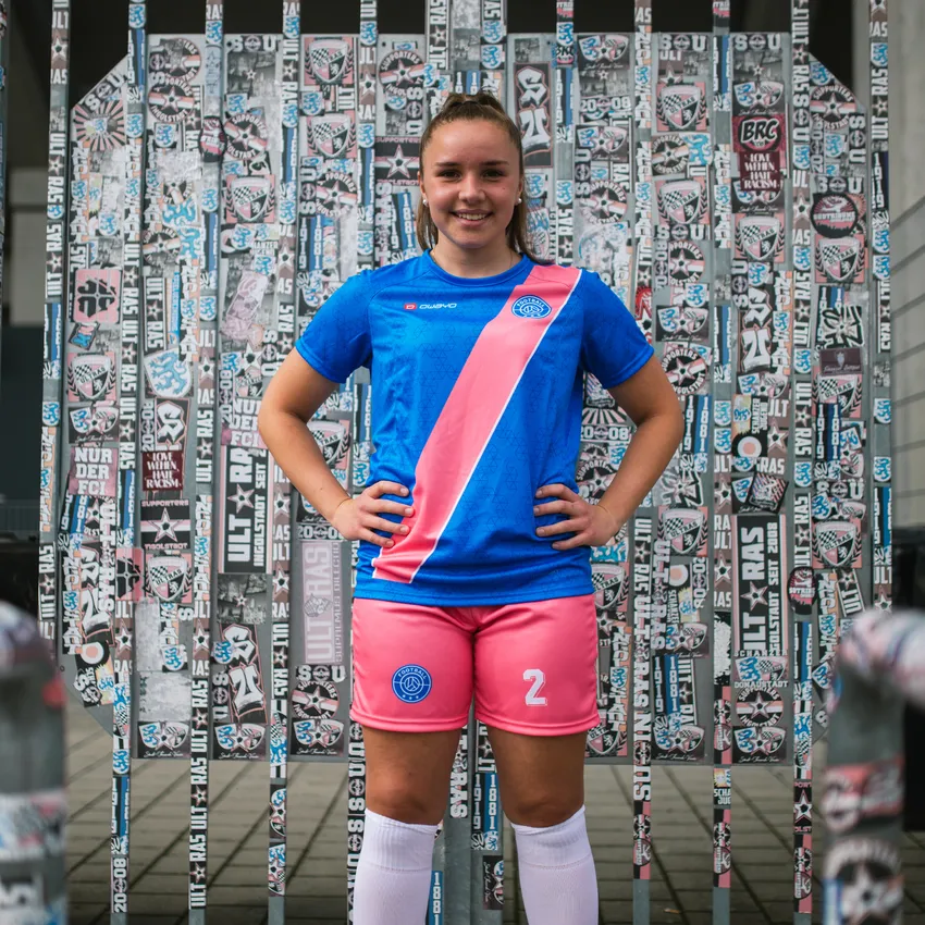 Una giovane ragazza in una maglietta da calcio blu con una striscia rosa e pantaloni rosa si trova davanti a un muro ricoperto di adesivi.