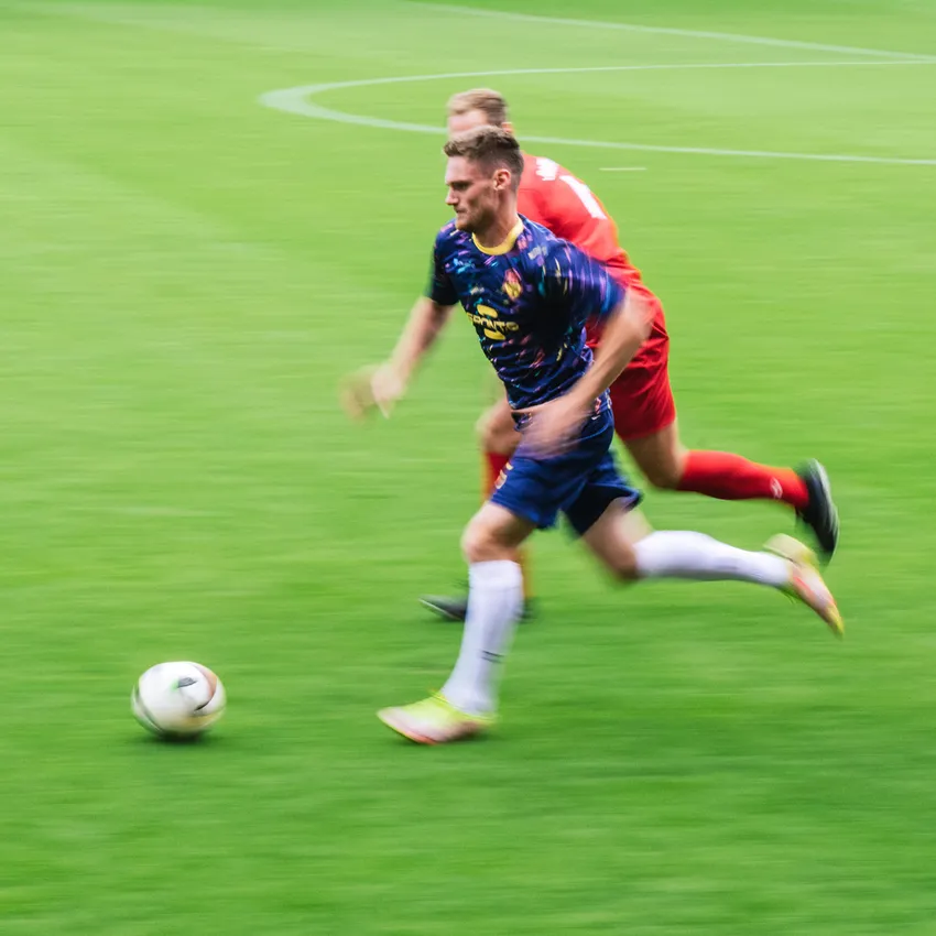 Un calciatore in una maglia colorata corre sul campo mentre un altro giocatore lo insegue.