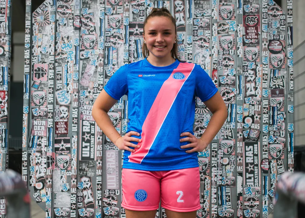 Una giovane atleta posa in una maglietta da calcio blu con una striscia rosa e pantaloni corti rosa davanti a un muro colorato coperto di adesivi.