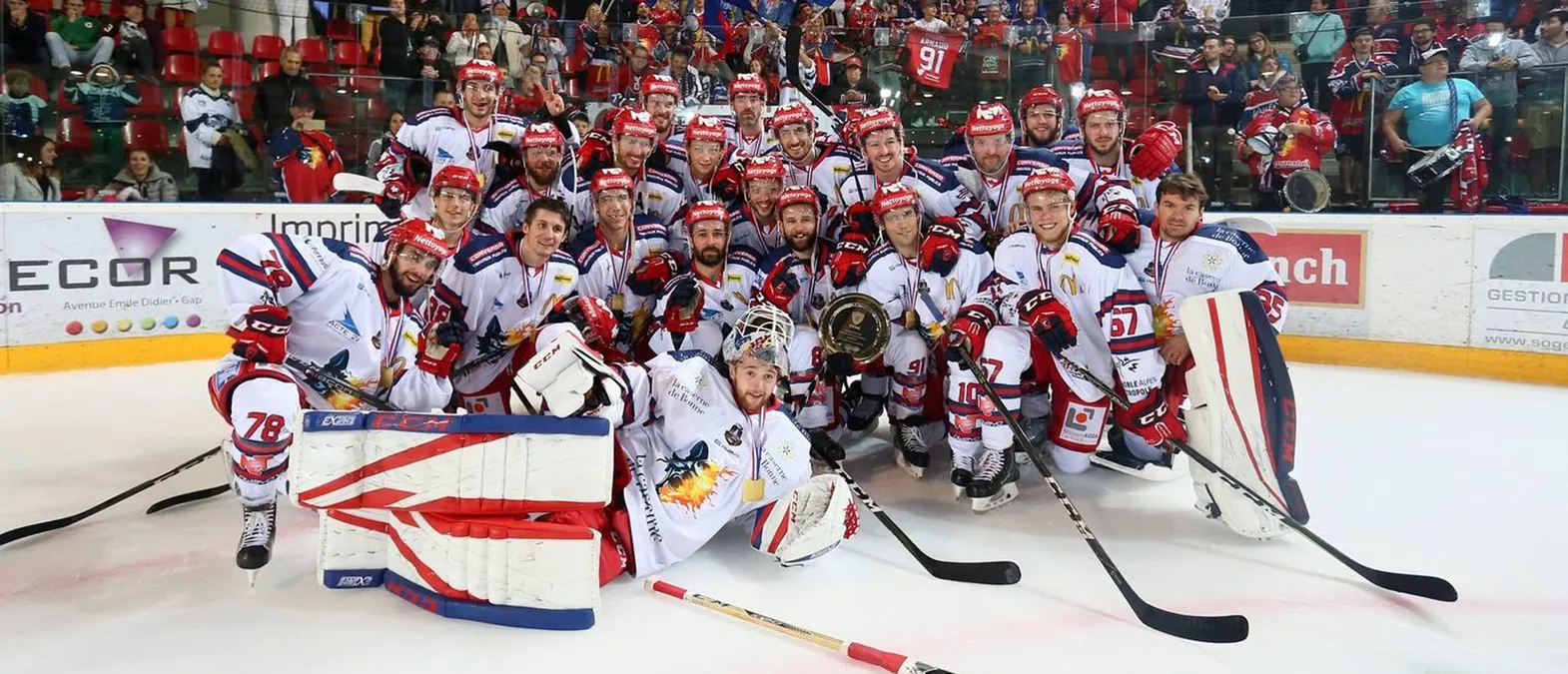 Una squadra di hockey su ghiaccio con maglie bianche con dettagli rossi e blu posa sul ghiaccio, circondata da tifosi, mentre i giocatori indossano medaglie e tengono un trofeo.
