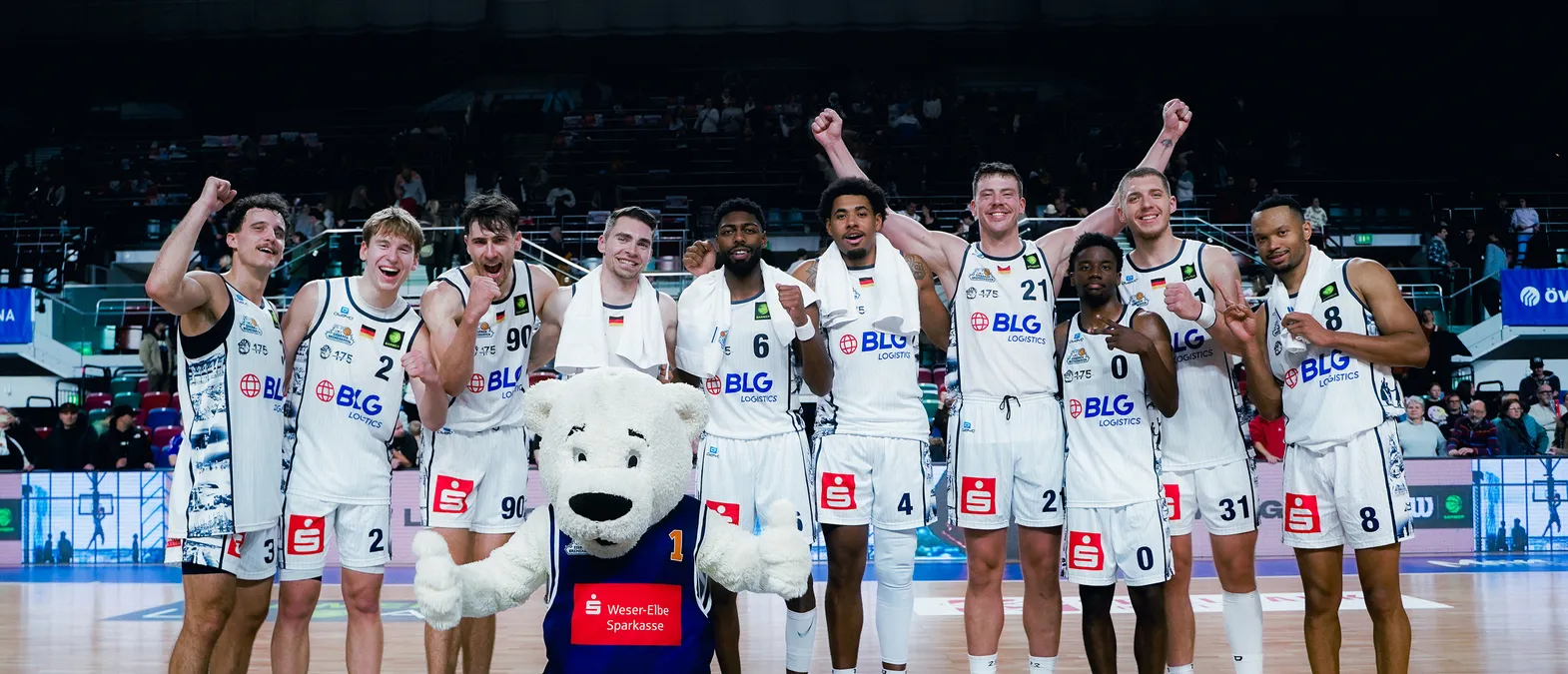 Giocatori di pallacanestro in maglie bianche con il logo di BLG Logistics festeggiano sul campo, circondati da un pubblico, con una mascotte orso polare in primo piano.