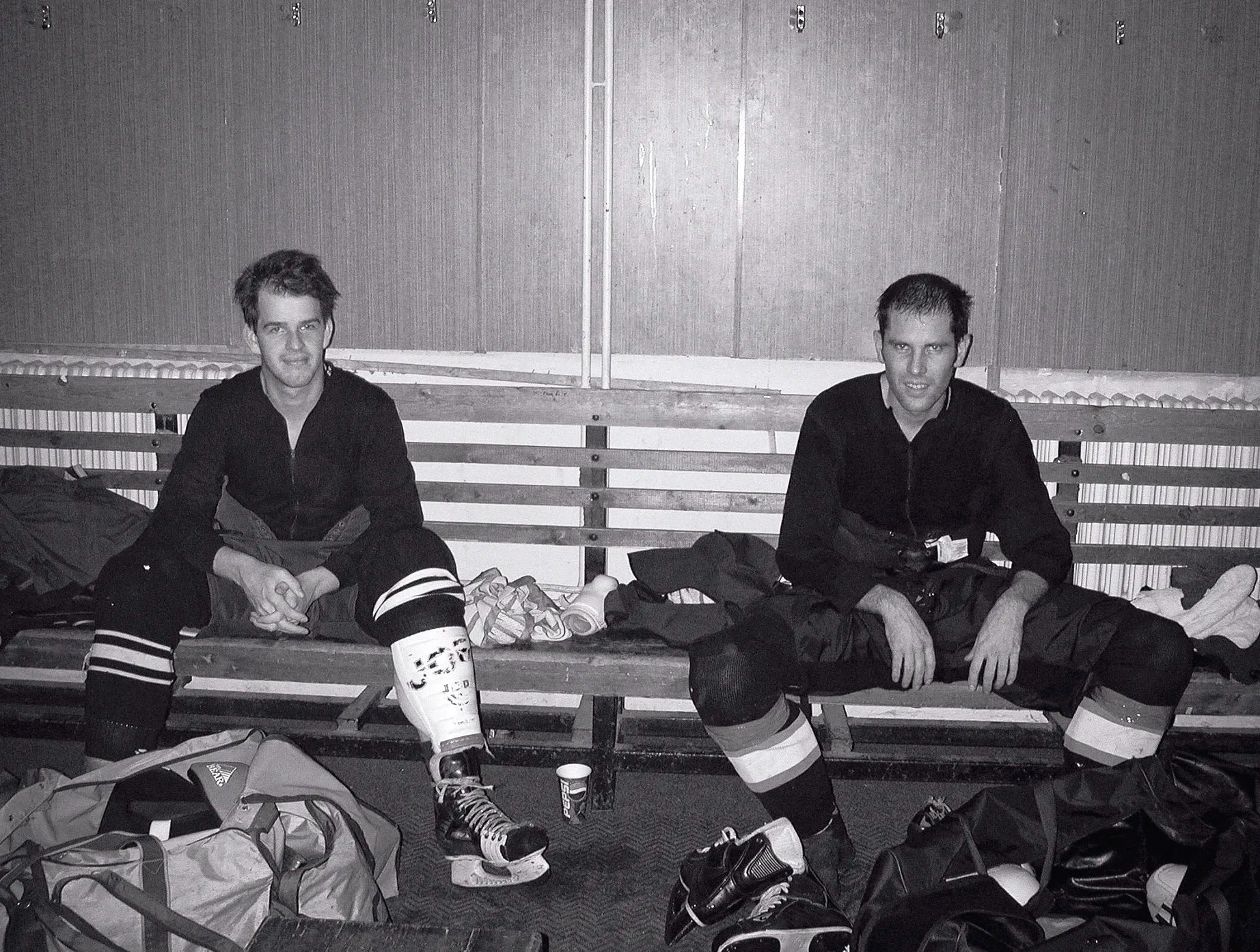 Due giocatori di hockey su ghiaccio in completa attrezzatura seduti su una panca di legno in un camerino, circondati da borse da sport e pattini.