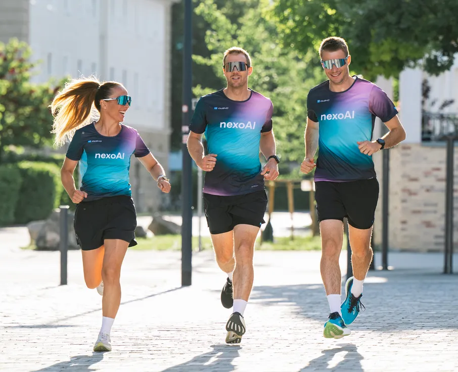 Tre atleti, due uomini e una donna, corrono indossando magliette da corsa colorate abbinate e pantaloni corti su un percorso pavimentato.