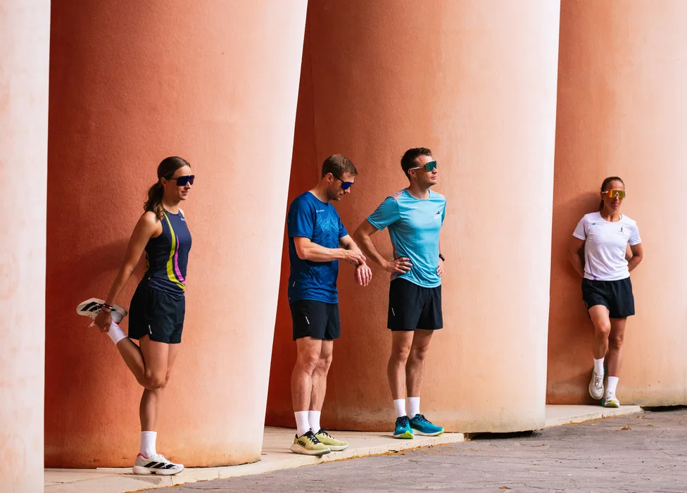 Un gruppo di quattro atleti in abbigliamento da corsa si sta allungando contro un muro con grandi colonne.