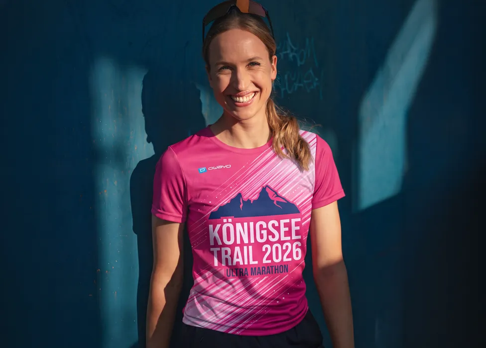 Una donna sorridente indossa una maglietta rosa con la stampa 'Königsee Trail 2026 Ultra Marathon' davanti a un muro blu.