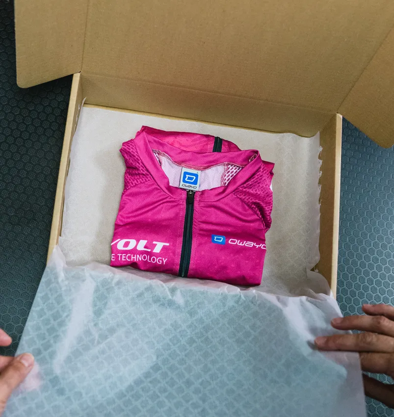 Un capo sportivo personalizzato rosa di owayo, piegato, è posizionato in una scatola di cartone aperta su carta bianca, tenuto da mani.