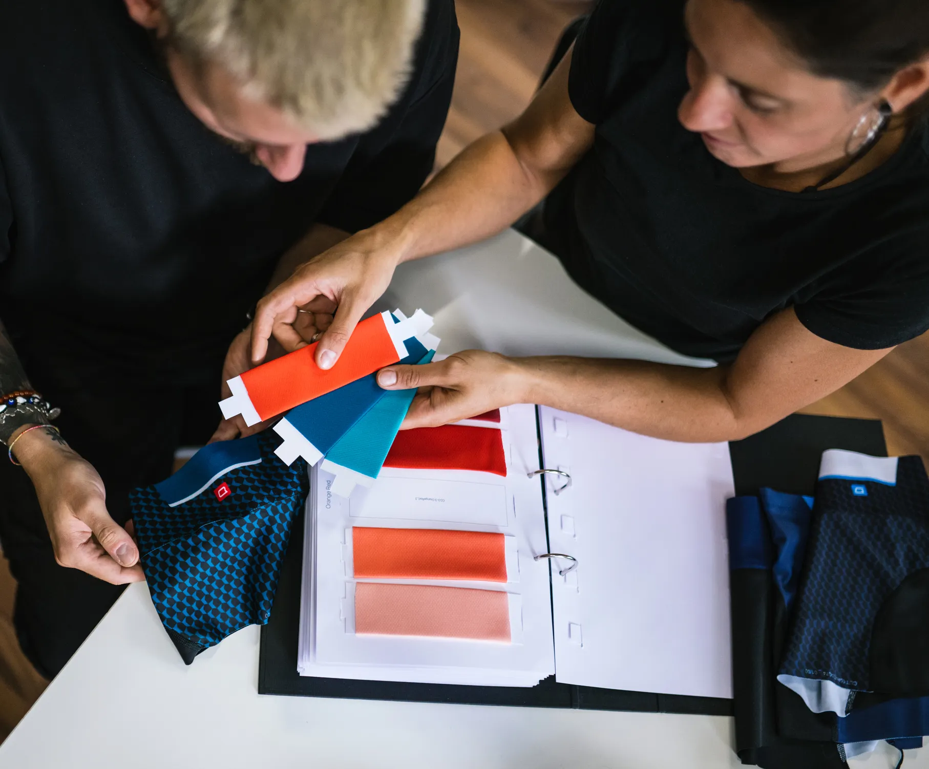 Due persone scelgono i colori per abbigliamento sportivo su misura esaminando campioni di tessuto e un catalogo colori.