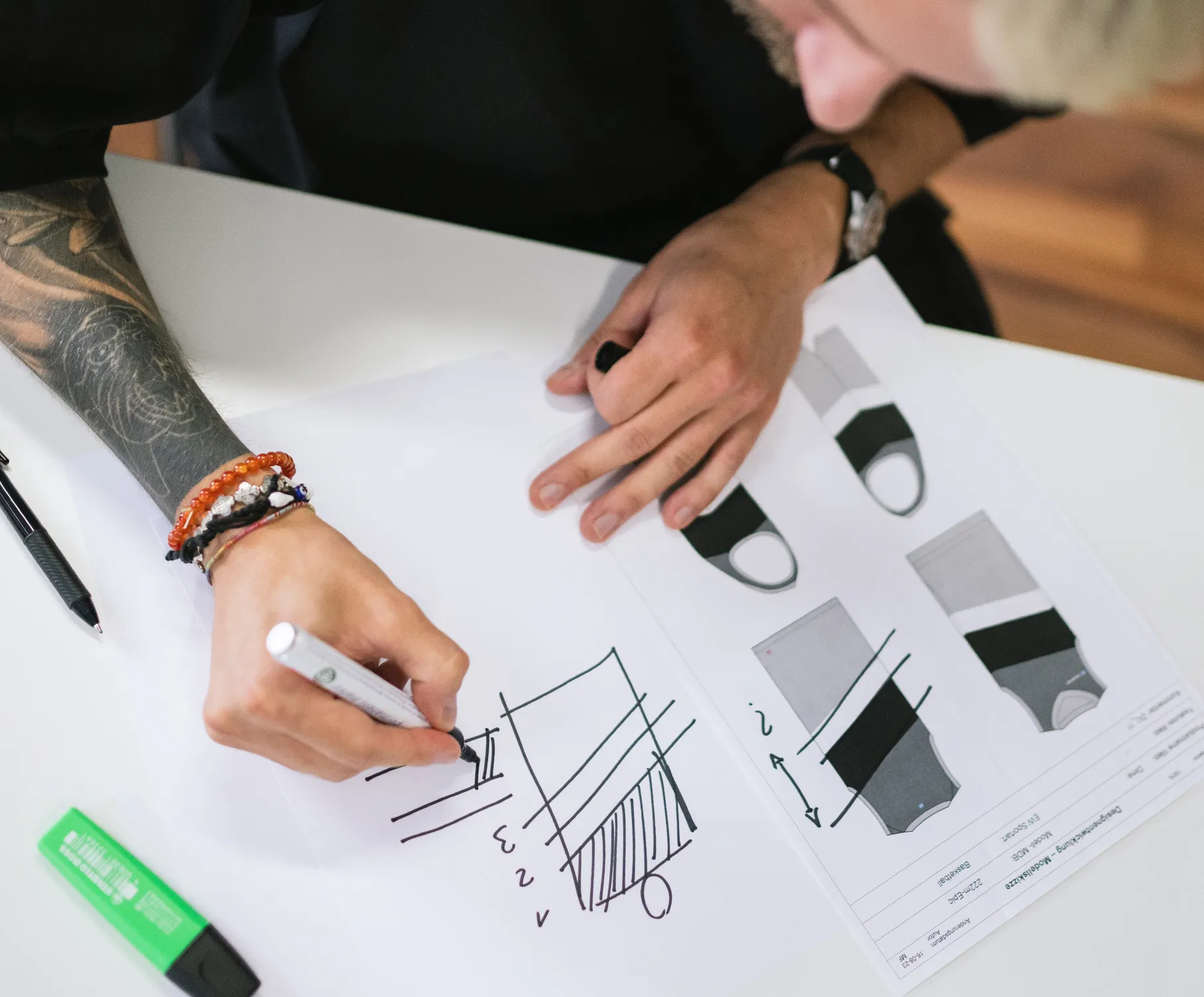 Una persona con un tatuaggio sul braccio disegna con un pennarello su un foglio di carta, mentre sul tavolo ci sono disegni tecnici di abbigliamento sportivo.