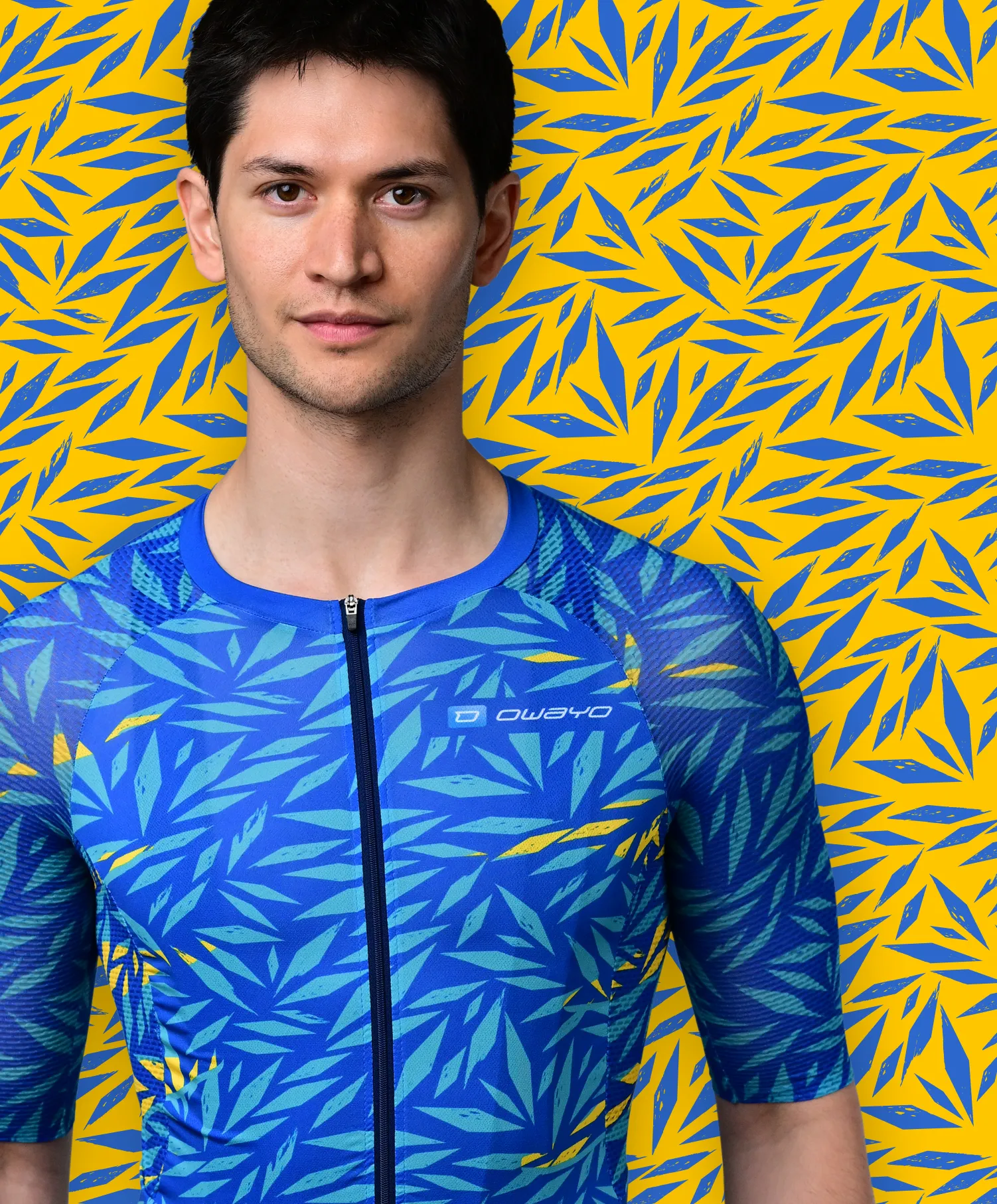 Un uomo indossa una maglia sportiva blu a maniche corte con un motivo di foglie blu e gialle che ricorda una pianta tropicale, davanti a uno sfondo giallo con motivi di foglie blu.