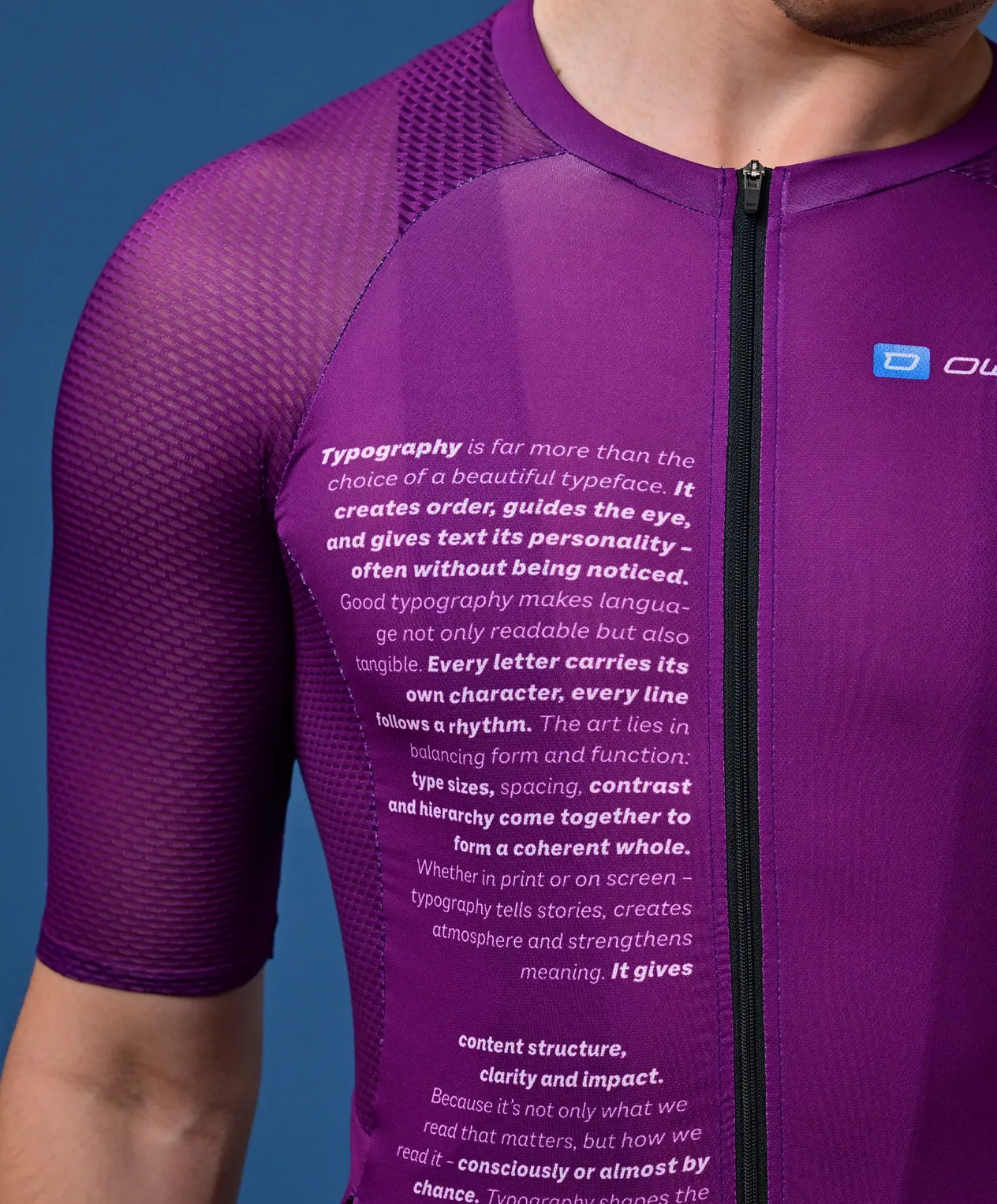 Un uomo indossa una maglia sportiva viola a maniche corte con un testo sulla tipografia sul petto e un piccolo logo sulla spalla.