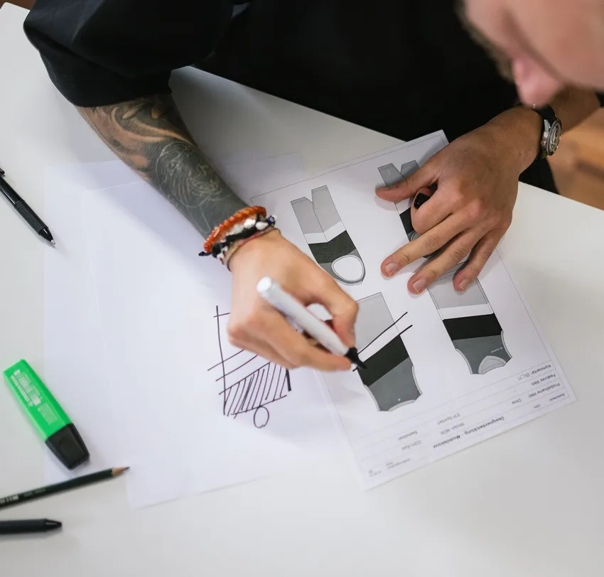 Una persona con un braccio tatuato disegna con un pennarello su un foglio di carta progetti per abbigliamento sportivo personalizzato.