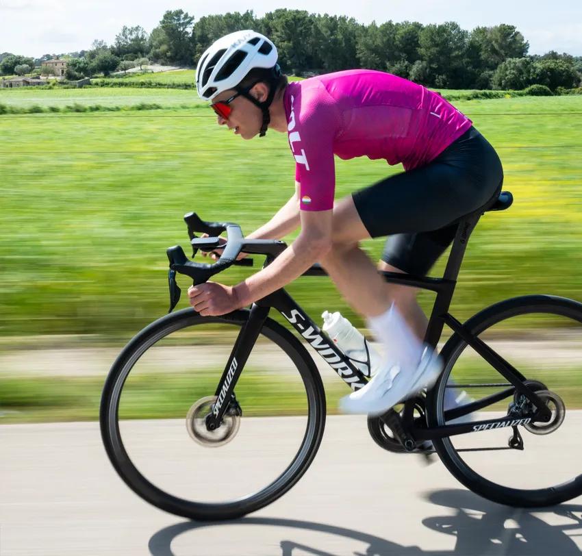 Un ciclista in una maglia rosa e pantaloncini neri pedala su una strada attraverso un paesaggio verde, con uno sfondo sfocato che enfatizza la velocità.