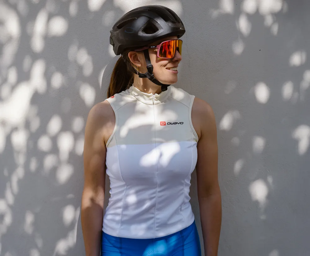 Una donna con una maglia da ciclismo bianca senza maniche con il logo owayo, un casco da bicicletta e occhiali da sole, appoggiata a un muro con ombre di foglie.