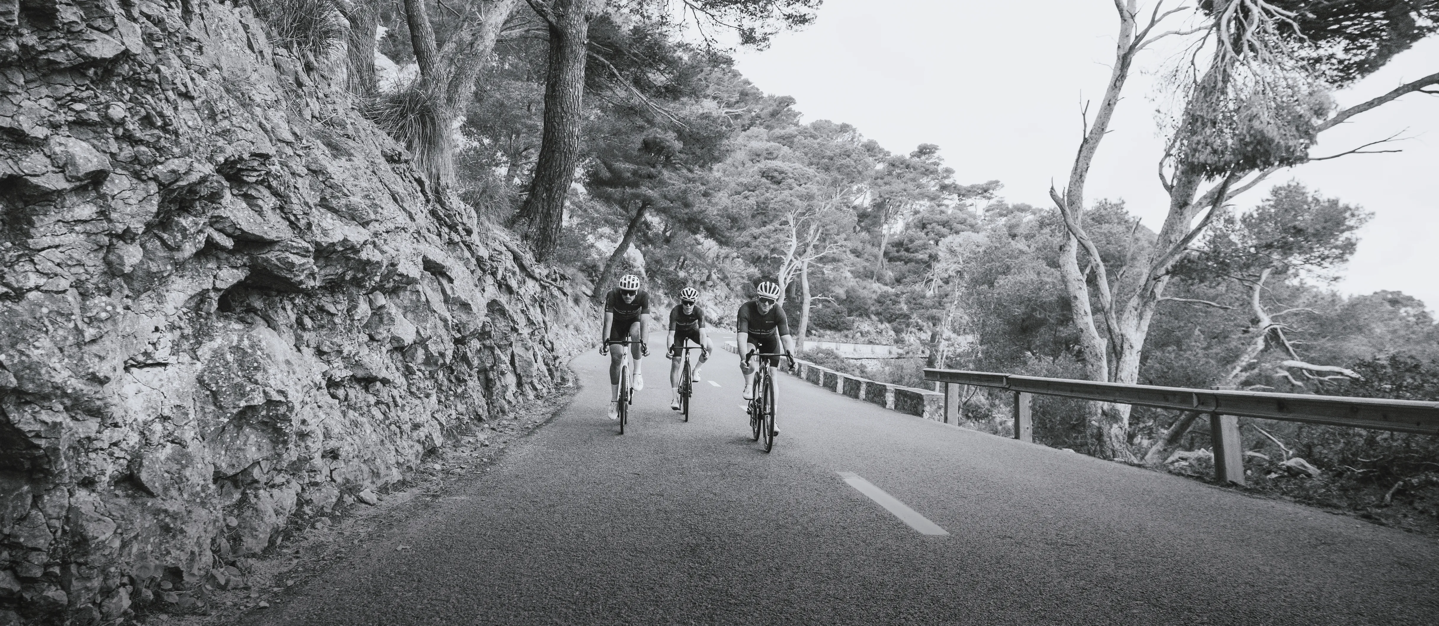 Tre ciclisti in abbigliamento sportivo pedalano su una strada lungo una scogliera rocciosa.
