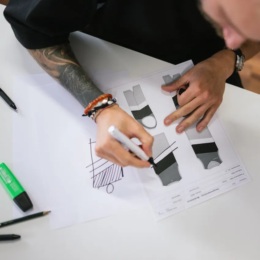 Una persona con un braccio tatuato sta disegnando progetti per abbigliamento sportivo su carta, circondata da strumenti di scrittura.