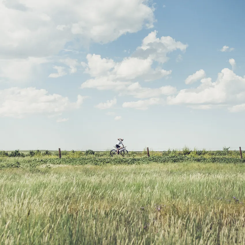 Una persona pedala su una bicicletta attraverso un paesaggio verde sotto un cielo blu con alcune nuvole.
