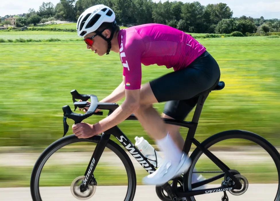 Un ciclista in una maglia rosa e pantaloncini neri pedala su una strada attraverso un paesaggio verde, con lo sfocato del movimento che enfatizza la velocità.