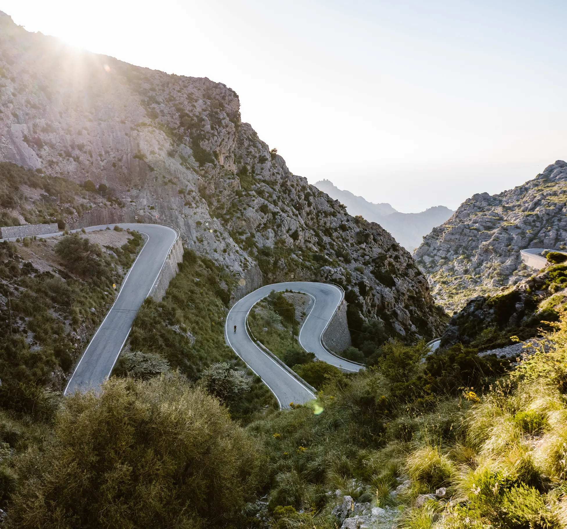 Una persona guida una bicicletta lungo una strada di montagna tortuosa che serpeggia attraverso un paesaggio roccioso, con la luce del sole che splende dall'alto.