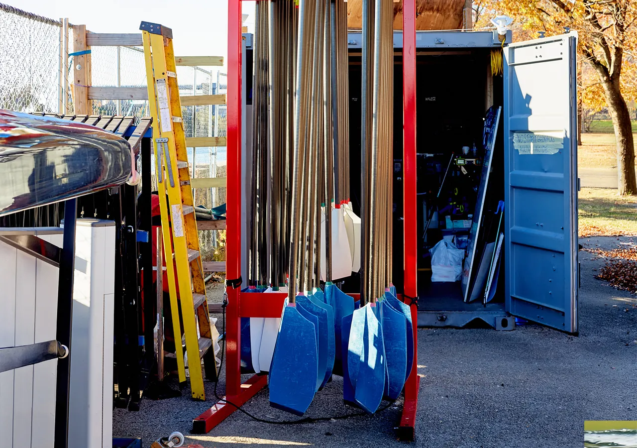 Un'immagine di un'area di stoccaggio di barche con remi di vari colori, una scala gialla e un container aperto sullo sfondo.