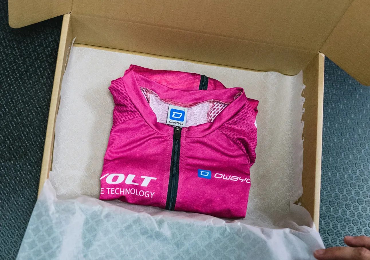 Una maglia sportiva rosa senza maniche piegata con il logo owayo è posizionata in una scatola di cartone su carta bianca.