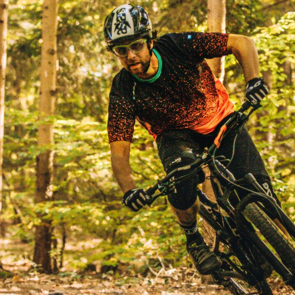 Un ciclista di mountain bike con una maglia nera con puntini rossi e pantaloncini neri pedala in un bosco.