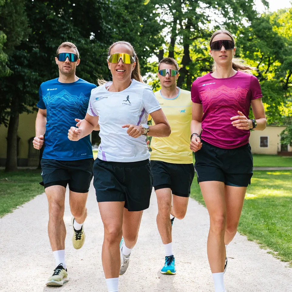 Quattro corridori in magliette sportive colorate e pantaloni corti corrono su un sentiero in un parco.