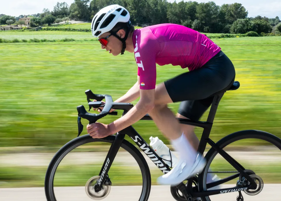 Un ciclista in una maglia rosa e pantaloncini neri pedala su una bicicletta da strada su una strada di campagna, con uno sfondo verde sfocato che suggerisce velocità.