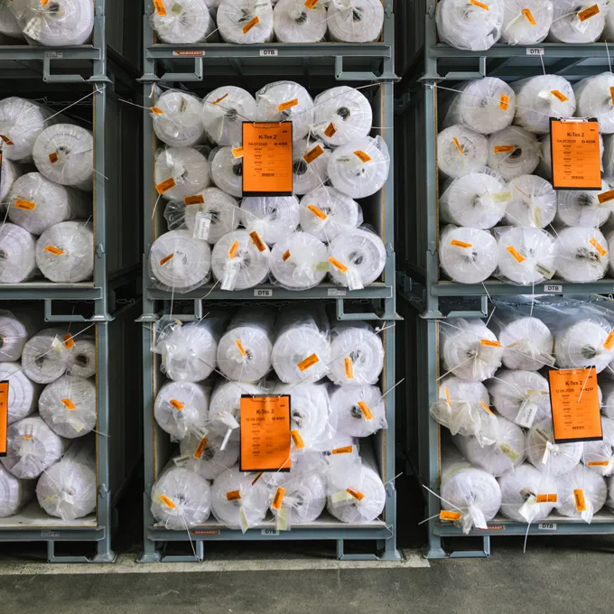 Un magazzino con scaffali pieni di grandi rotoli di tessuto bianco avvolti in plastica e contrassegnati con etichette arancioni.