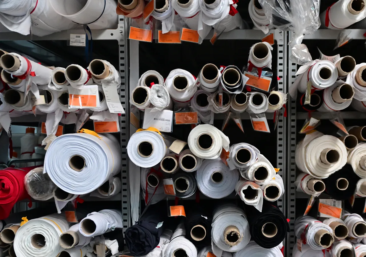 Scaffali pieni di rotoli di tessuto in vari colori, tra cui bianco, nero e rosso, con etichette che indicano una fabbrica di produzione interna.