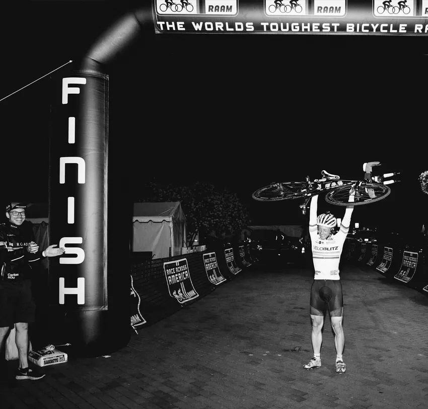 Un ciclista festeggia il traguardo della 'Race Across America' sollevando la bicicletta sopra la testa, mentre un uomo accanto a lui applaude.