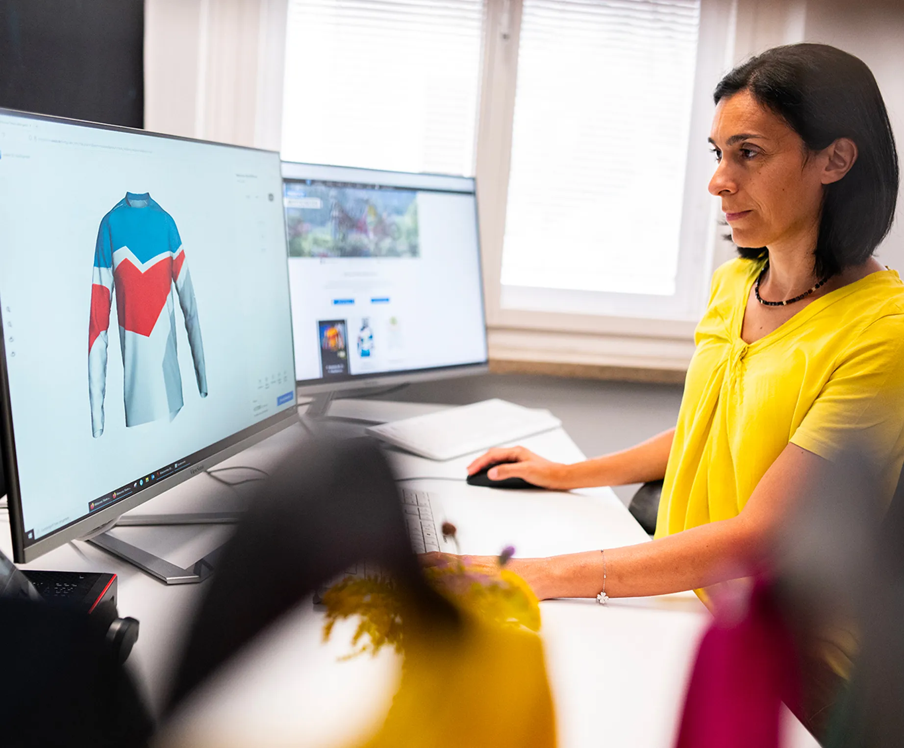 Una donna in una maglietta gialla è seduta a una scrivania e progetta su uno schermo del computer una maglia sportiva personalizzata con pannelli blu, rossi e bianchi.